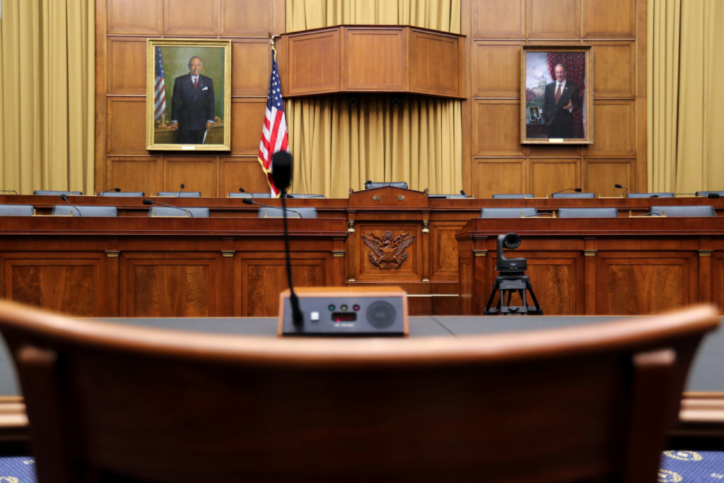 A real photograph of a congressional committee hearing room with lawmakers seated on the dais and a witness seated at a table with a microphone, news photography style