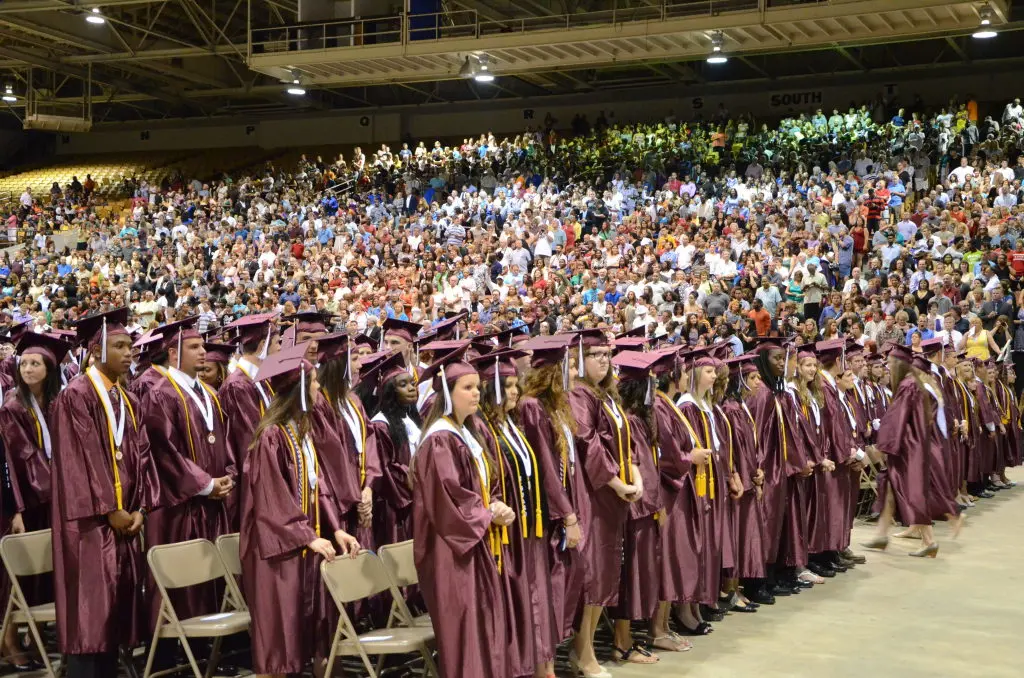 A real photograph of a high school graduation ceremony in the United States with graduates in caps and gowns seated while families watch from the audience