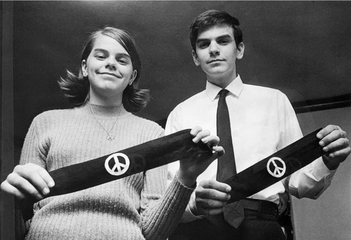 A real photograph of a high school student seated at a classroom desk wearing a plain armband on their sleeve, with a teacher blurred in the background
