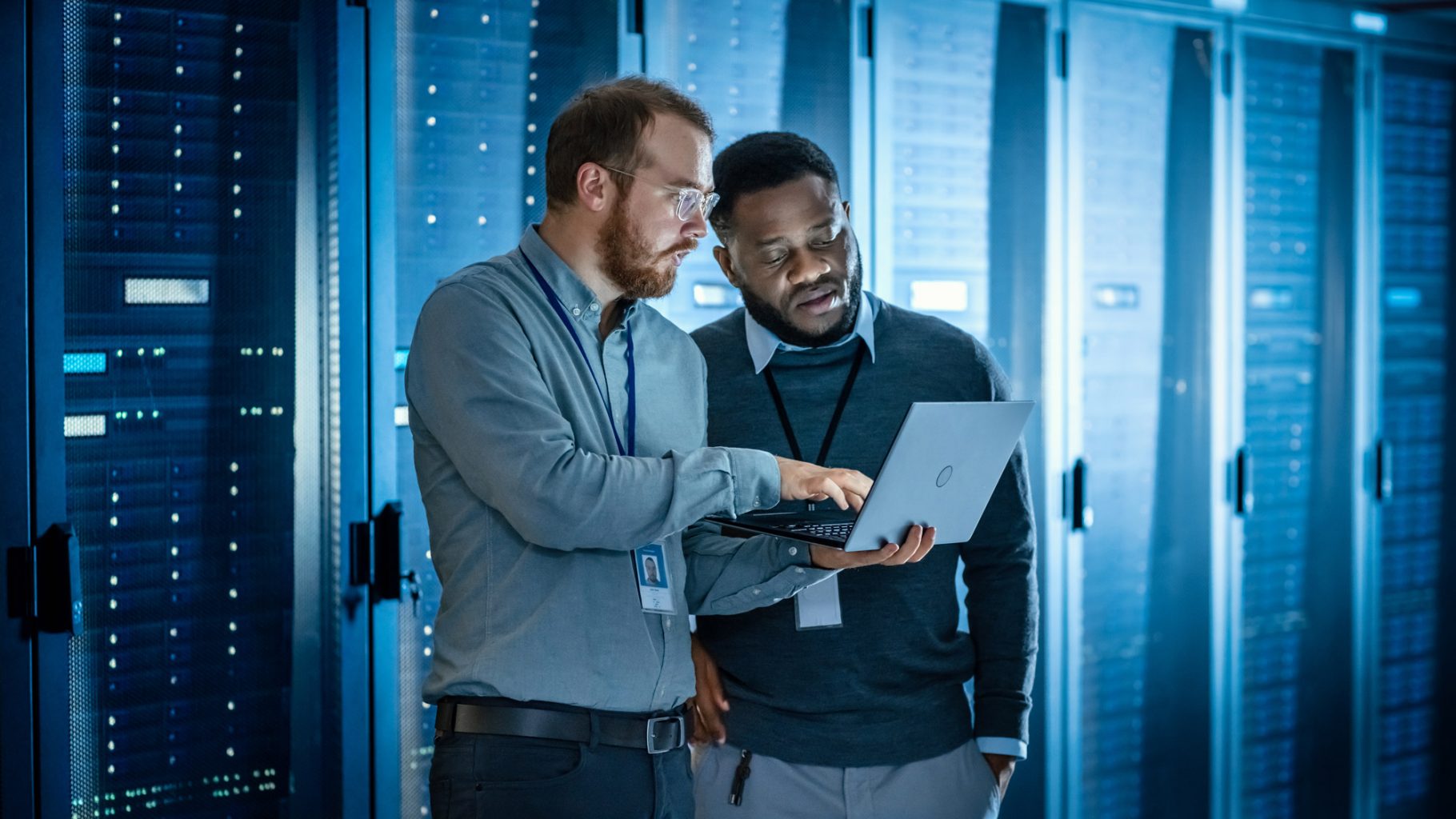 A real photograph of a technician in a telecommunications data center standing beside server racks and network equipment under bright industrial lighting