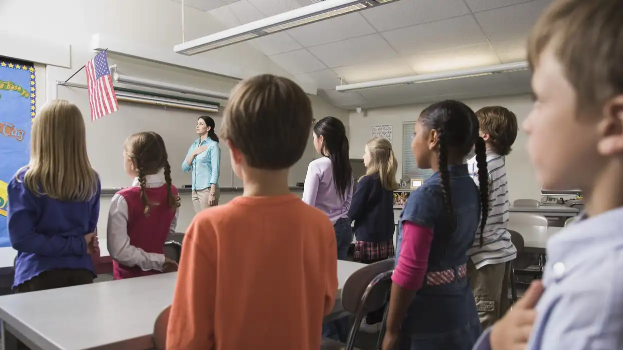 A real photograph of students in a civics classroom raising their hands while a teacher stands near a whiteboard, daytime light through classroom windows