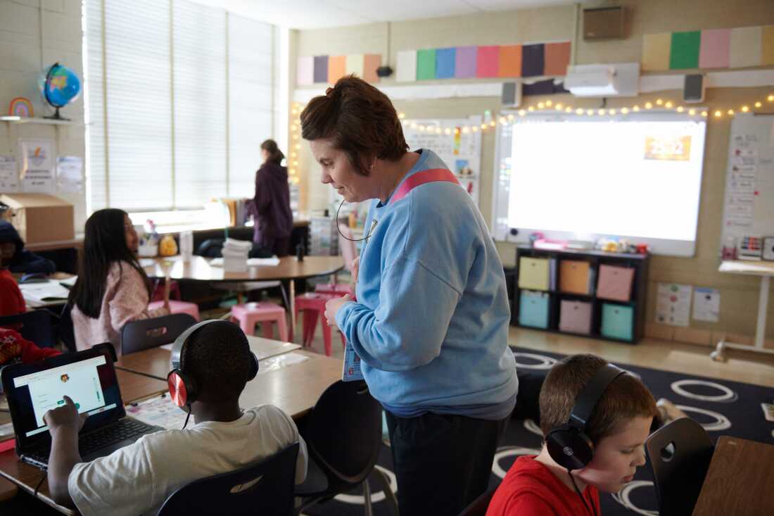 A real photograph of students in a journalism classroom working at computers with newspaper pages on screens, teacher standing nearby