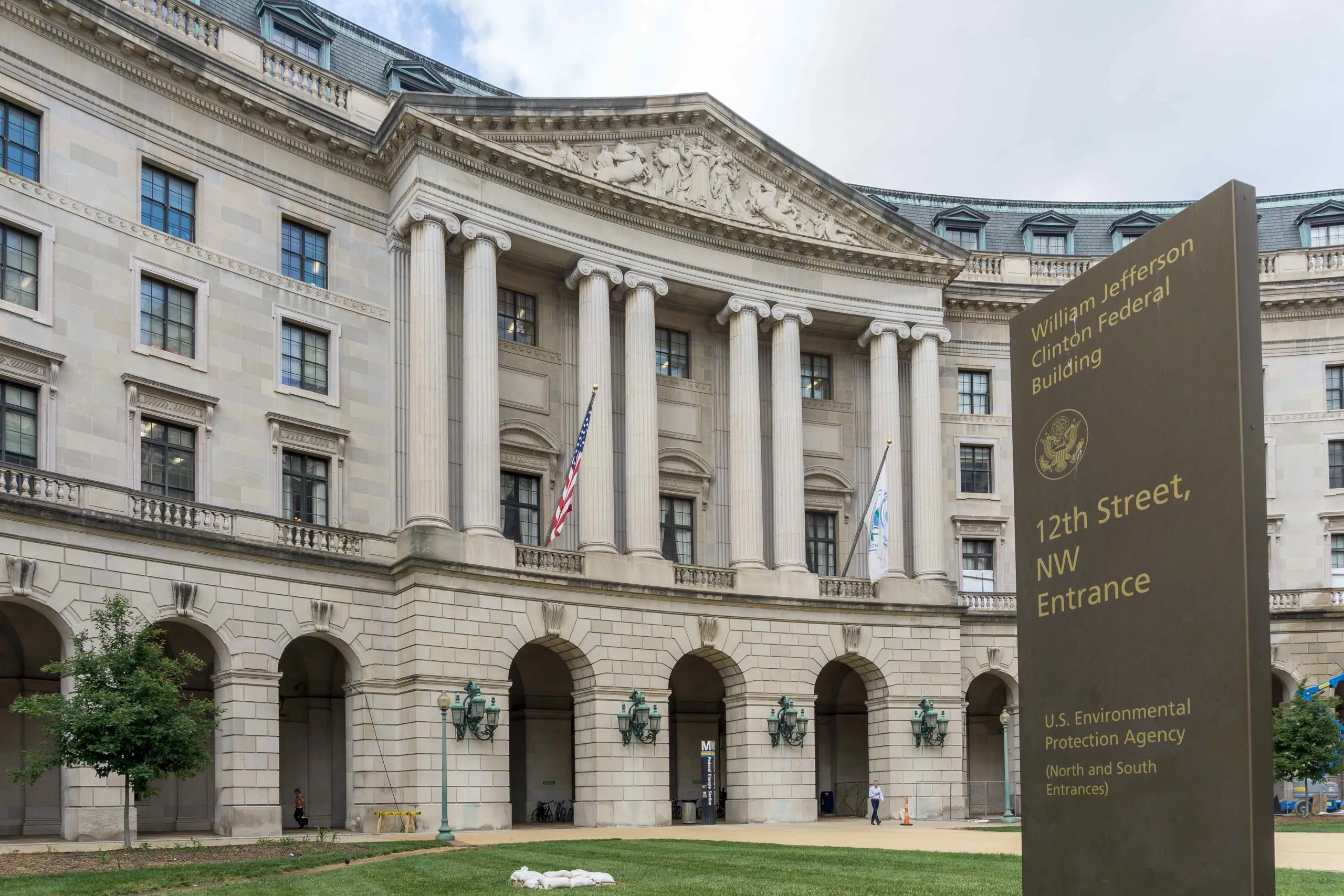 A real photograph of the Environmental Protection Agency headquarters building in Washington, D.C., with the EPA seal visible on the exterior wall and pedestrians passing by