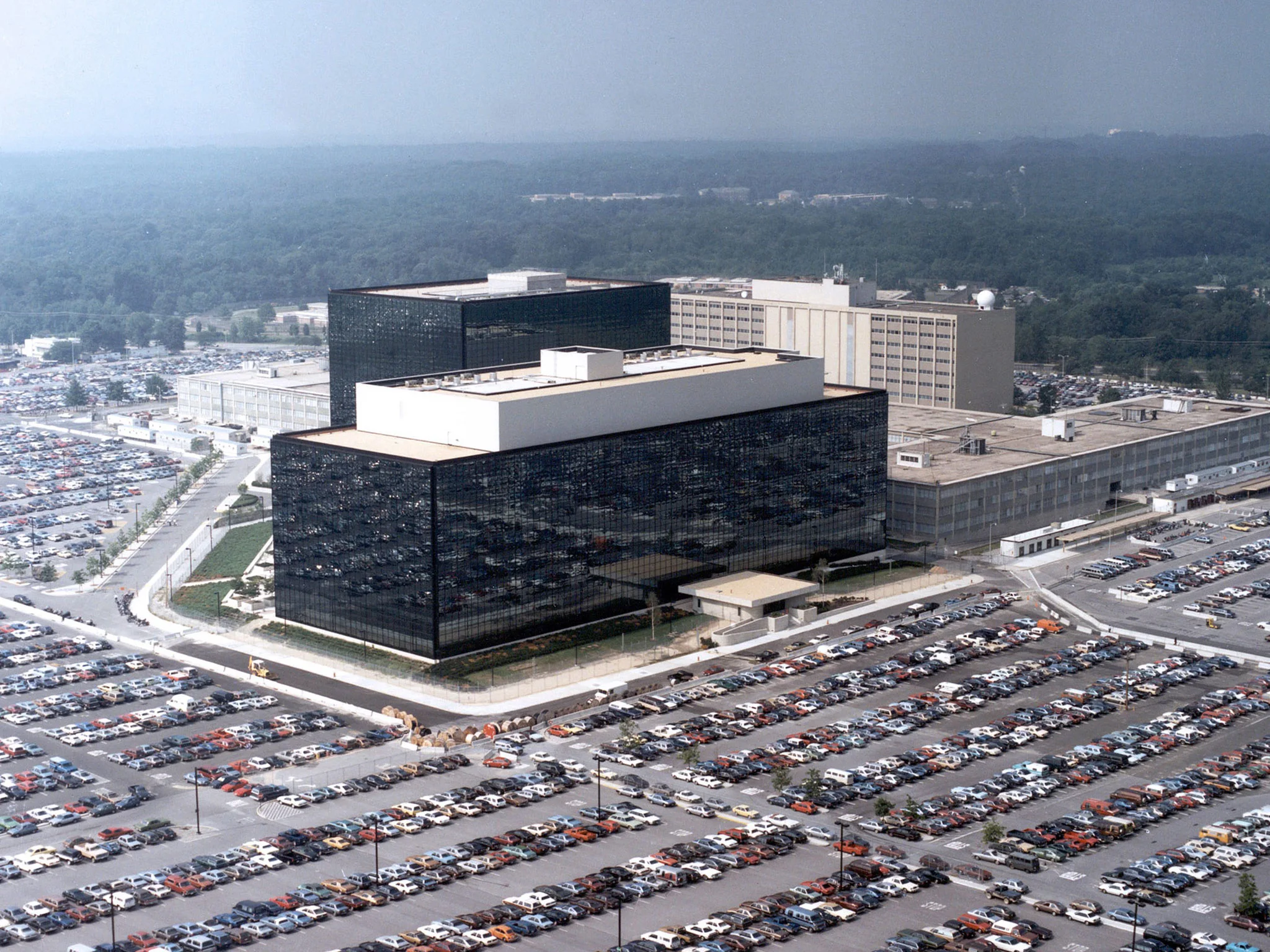 A real photograph of the National Security Agency headquarters at Fort Meade, Maryland on a clear day, showing the building exterior and security perimeter in a typical news photo style