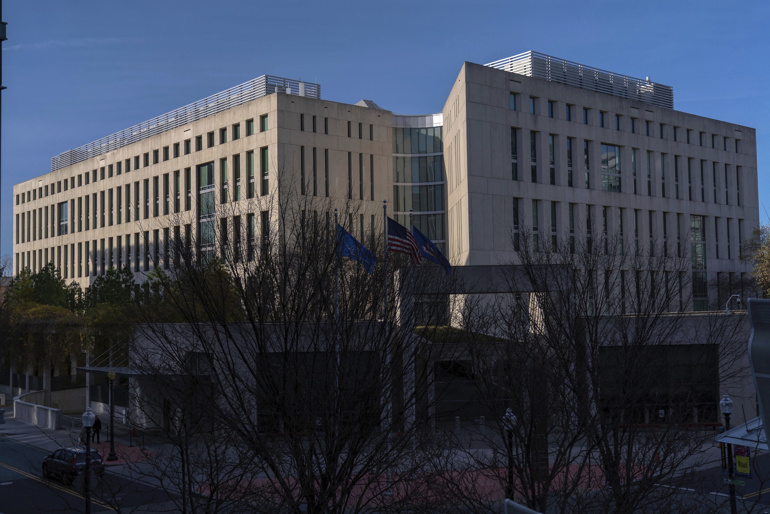 A real photograph of the Office of Management and Budget building exterior in Washington, DC on a clear day with pedestrians nearby