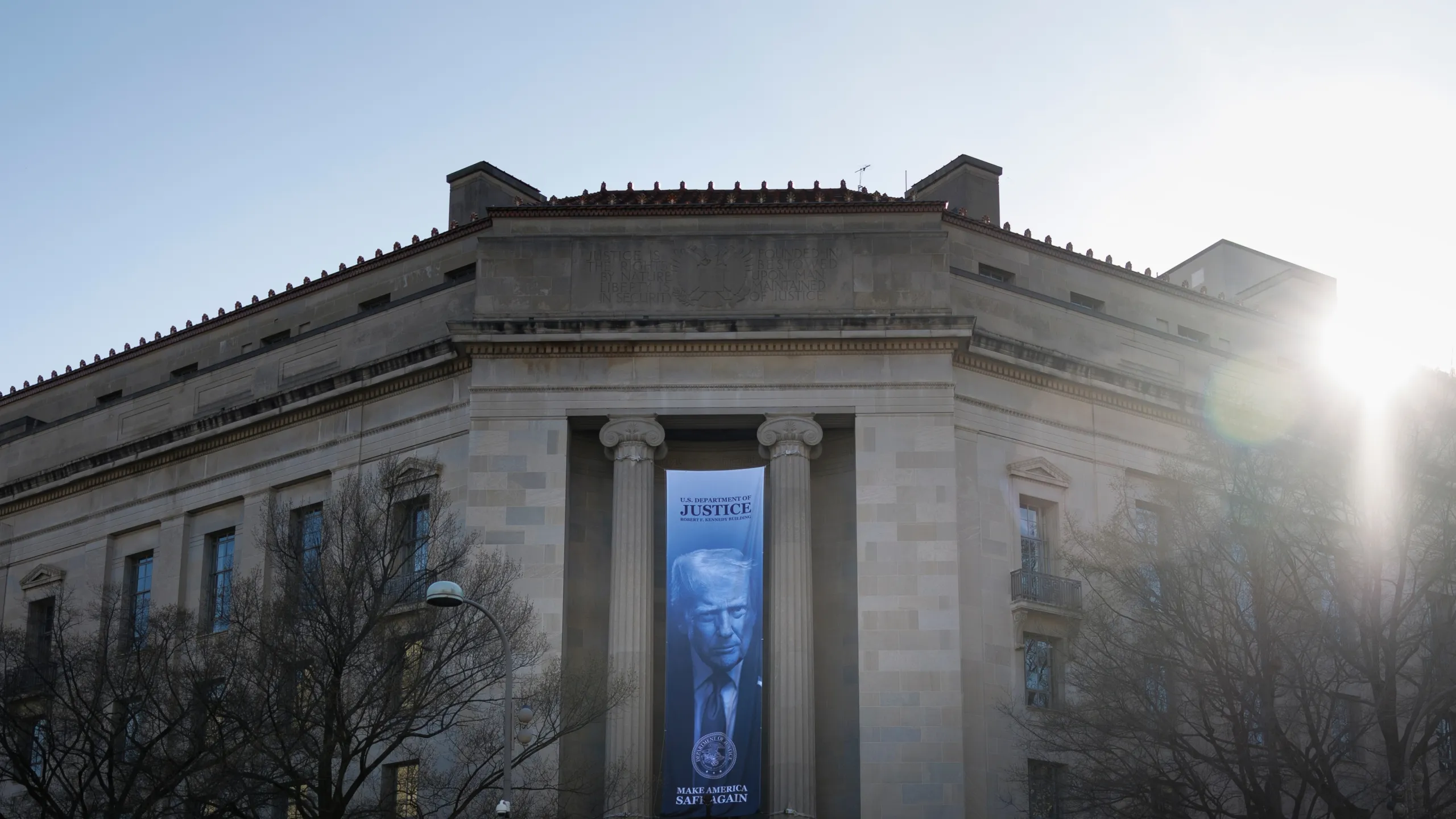 A real photograph of the exterior of the United States Department of Justice building in Washington, DC on a clear daytime sky with pedestrians in the distance