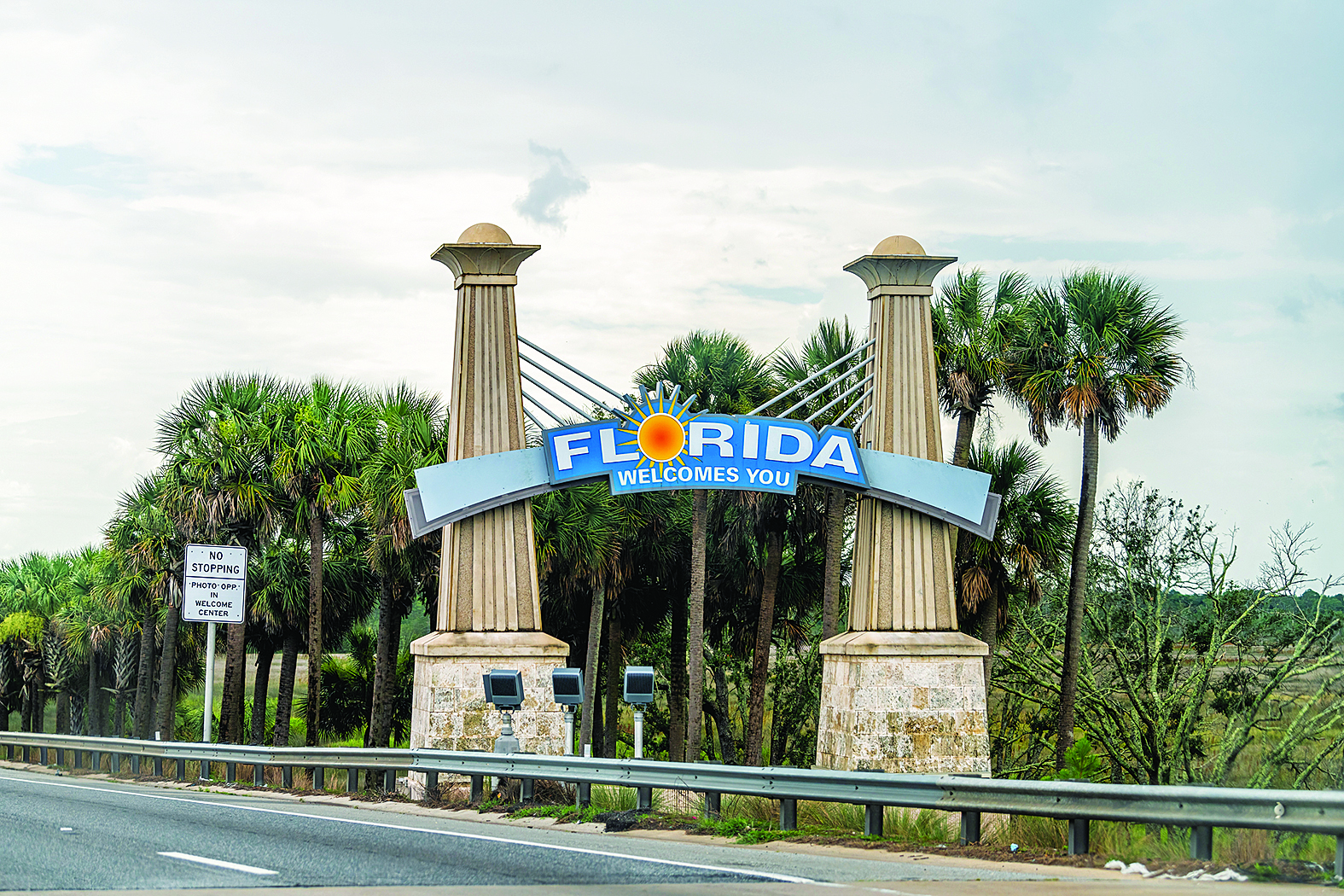 A real roadside welcome sign marking a state line on an interstate highway, with cars passing in the background in daylight, news photography style