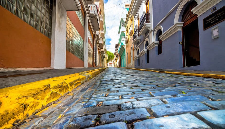 A real street scene in Old San Juan, Puerto Rico, with residents walking past historic buildings and an American flag visible in the background, documentary photography style