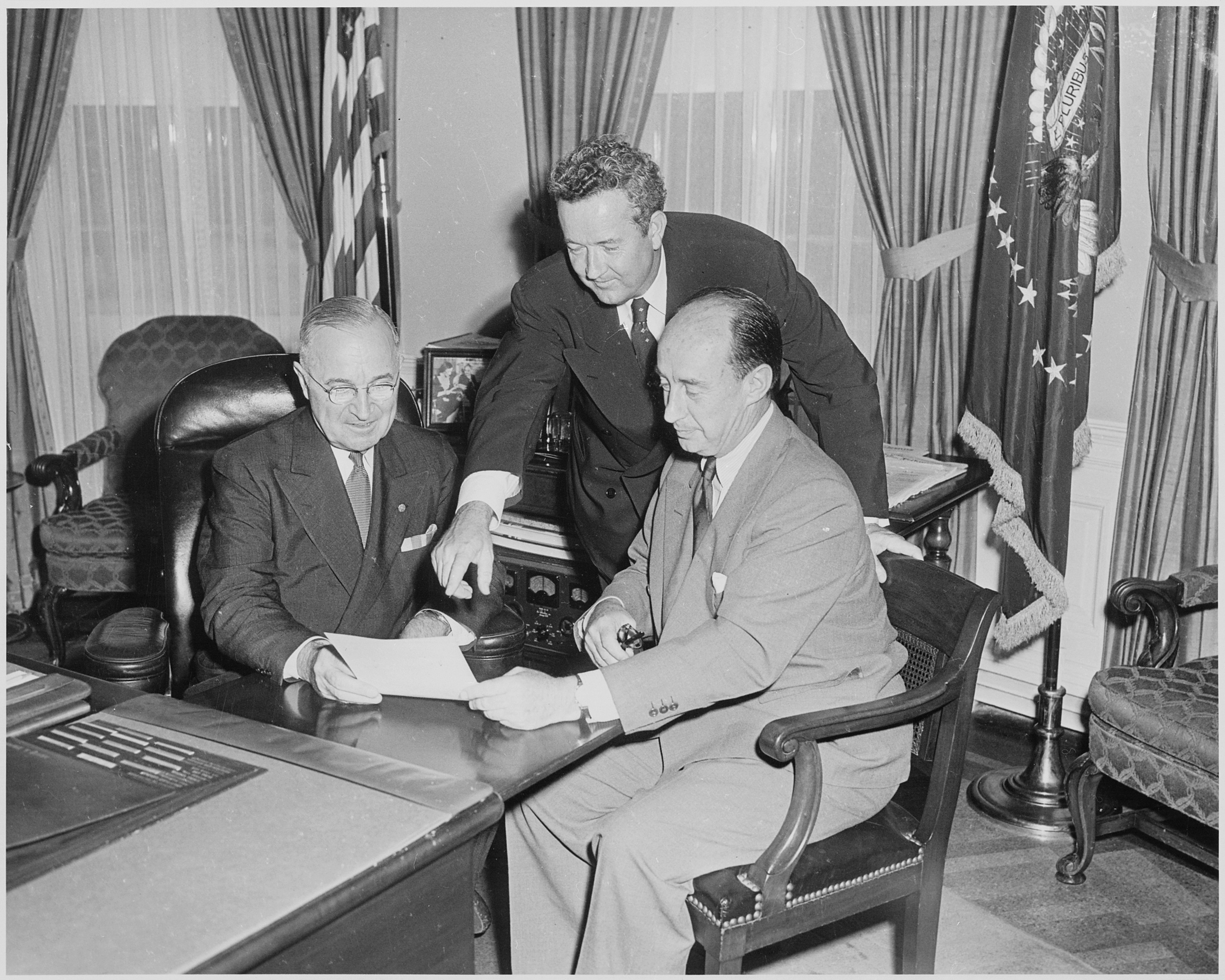 A realistic news-style photograph of President Harry S. Truman seated at a desk in the Oval Office, speaking to advisors during a tense policy moment, no visible text