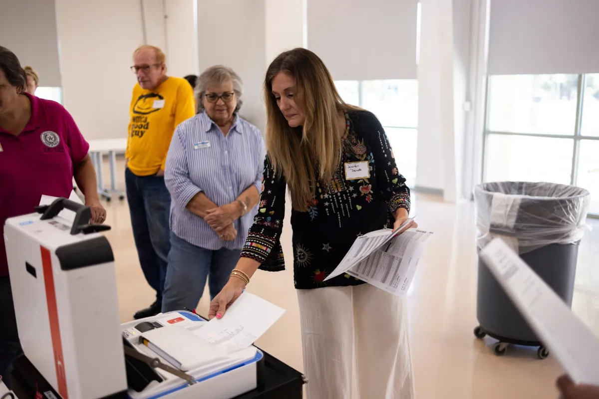 A realistic photograph of a county elections office public counter where a voter hands paperwork to an election clerk, with official office signage visible in the background but no readable text
