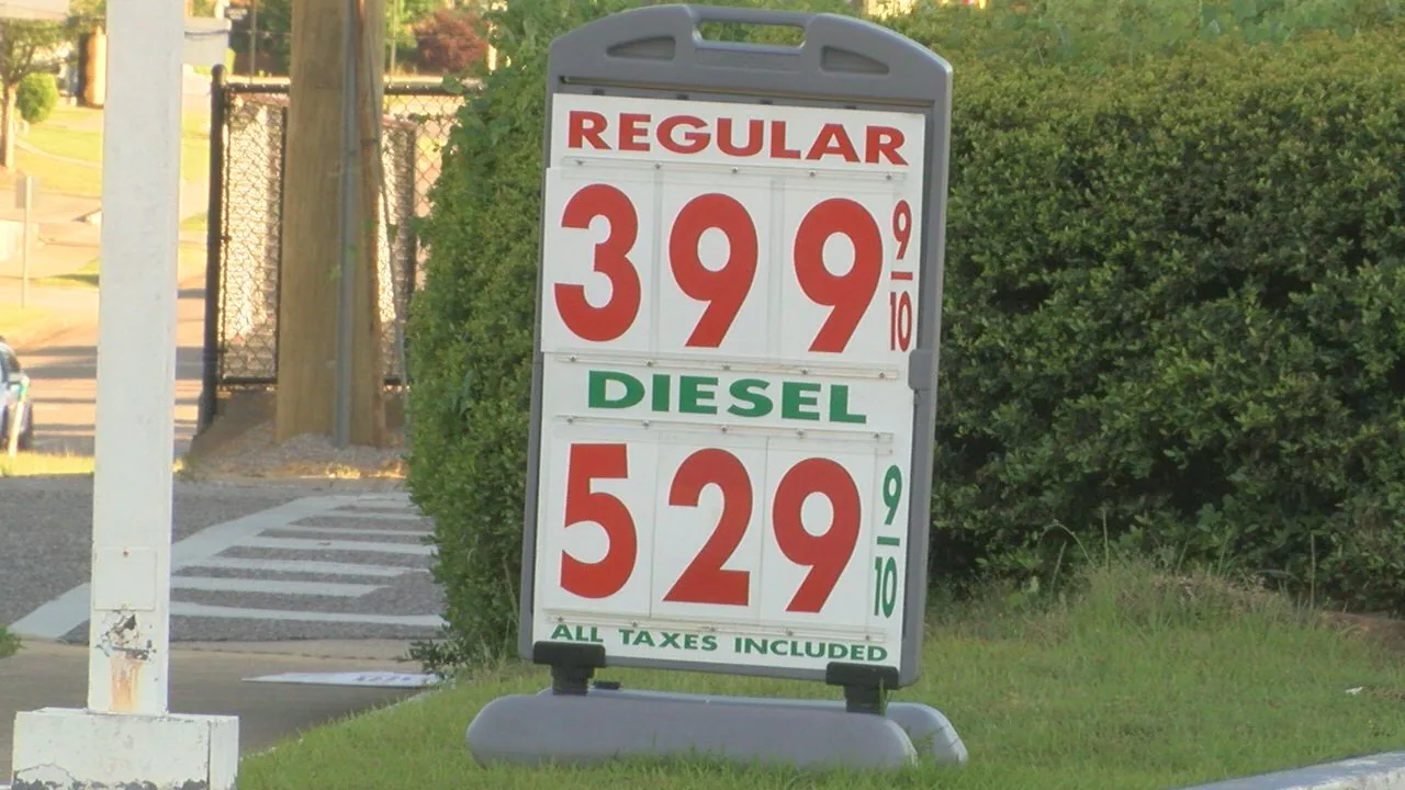 A roadside gas station price sign showing elevated gasoline and diesel prices, photographed at dusk with cars passing in the background, realistic news photography