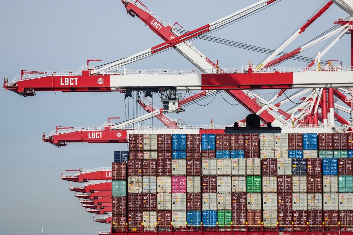 A row of stacked shipping containers at the Port of Los Angeles under overcast skies, with cranes in the background and trucks waiting to move freight, news photography style