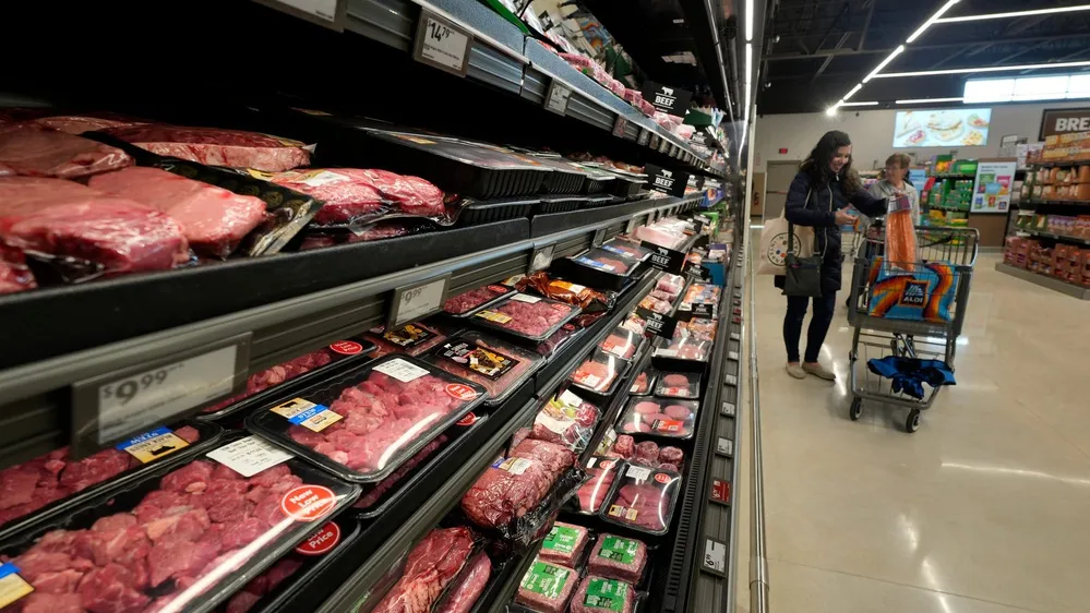A shopper pushing a cart down an American grocery store aisle with price tags visible on shelves, candid news photography style