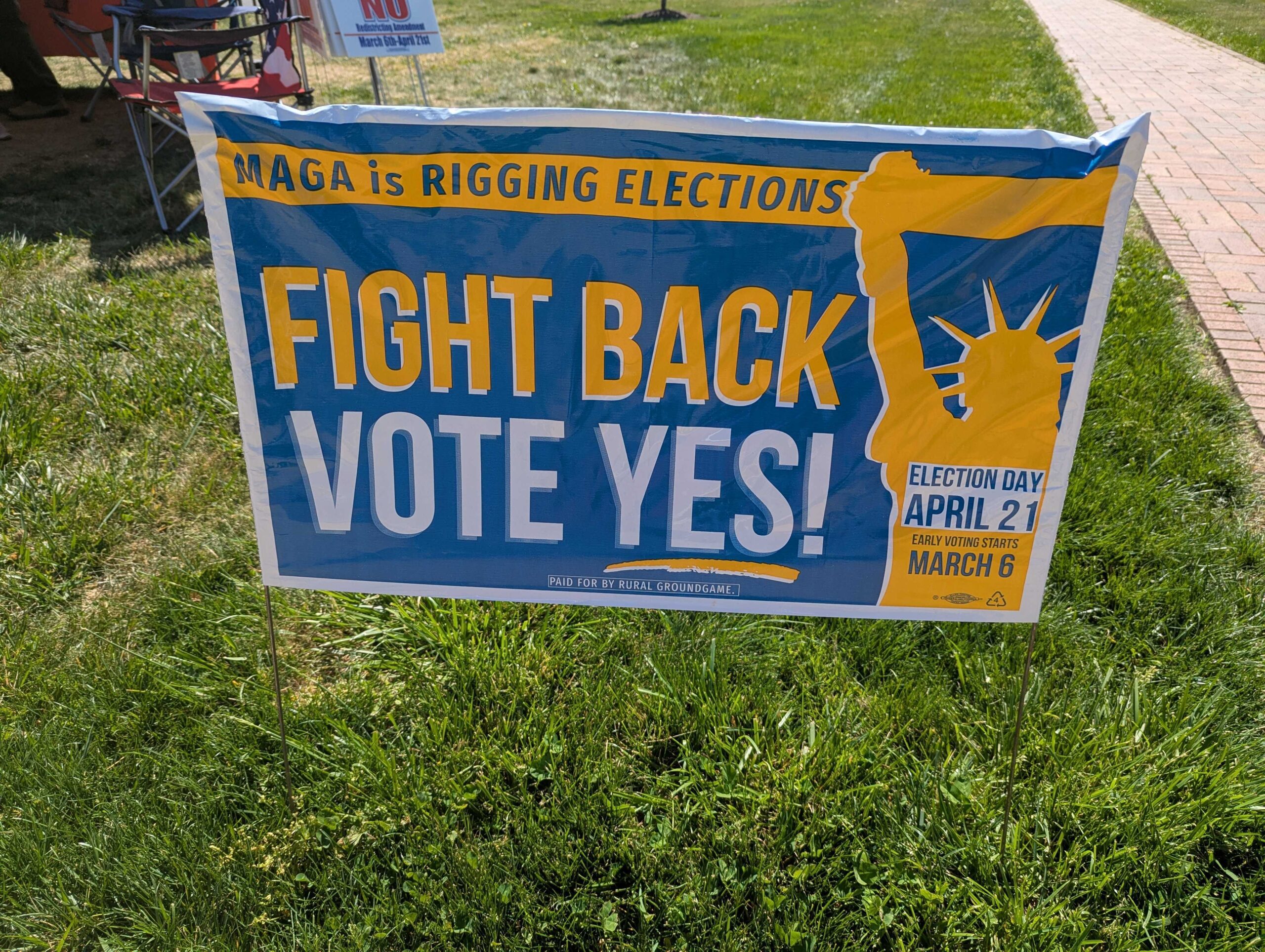 A single campaign yard sign planted on a lawn in a suburban American neighborhood during election season, candid news photography style