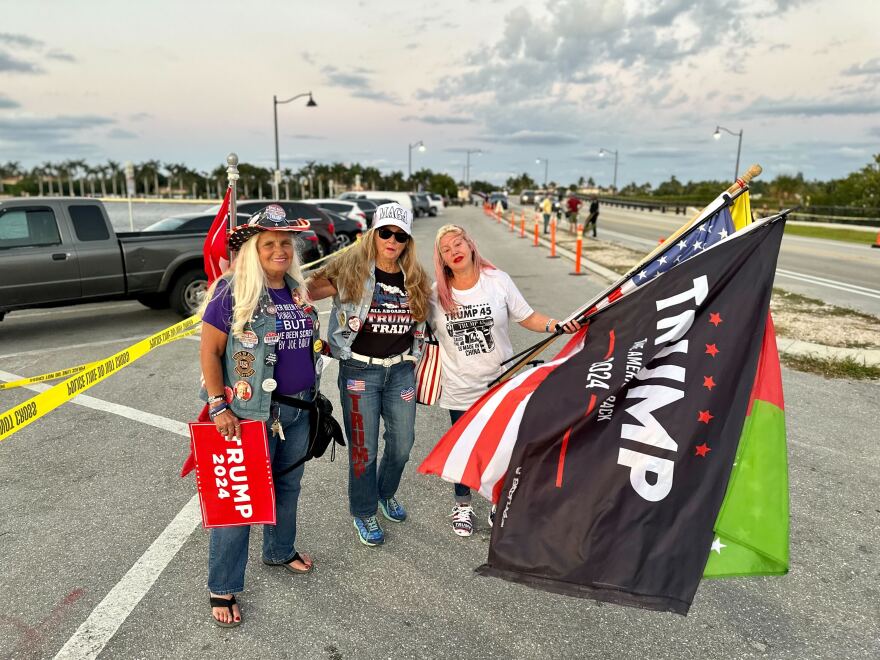A small group of supporters near Mar-a-Lago in Palm Beach, Florida, with a flag featuring Donald Trump waving in the foreground, news photography style
