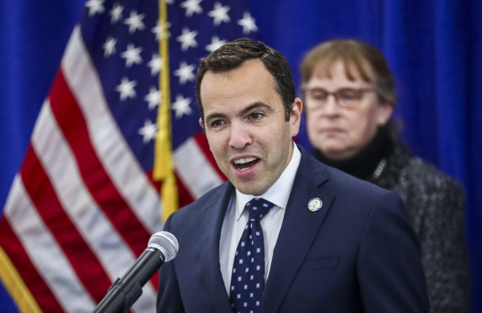 A state attorney general speaking at a podium during a press conference inside a government building, reporters and microphones in the foreground, news photography style