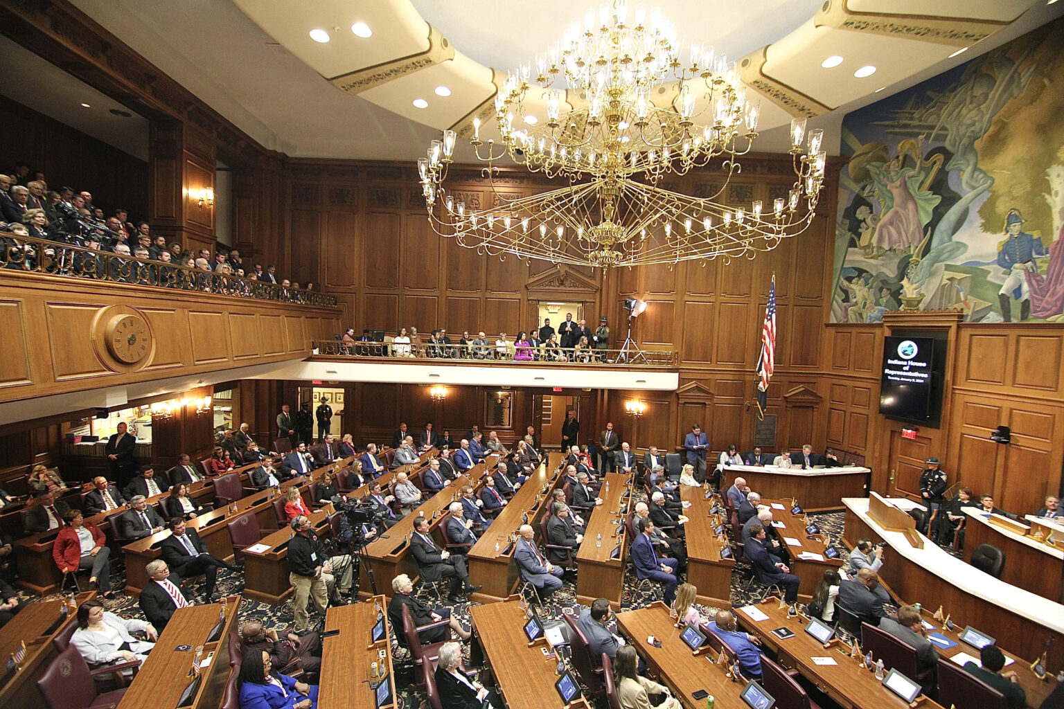 A state legislative chamber during a live session, lawmakers seated at desks with papers and microphones, candid news photography style