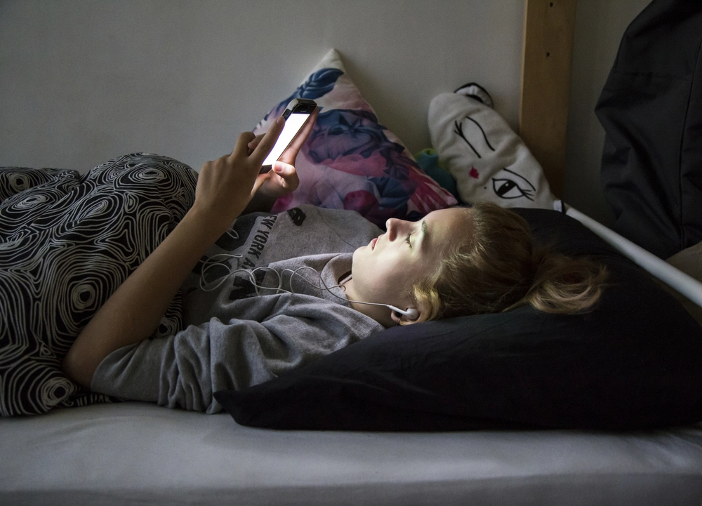 A teenager sitting up in bed at night lit by a smartphone screen in a dark bedroom, news photography style