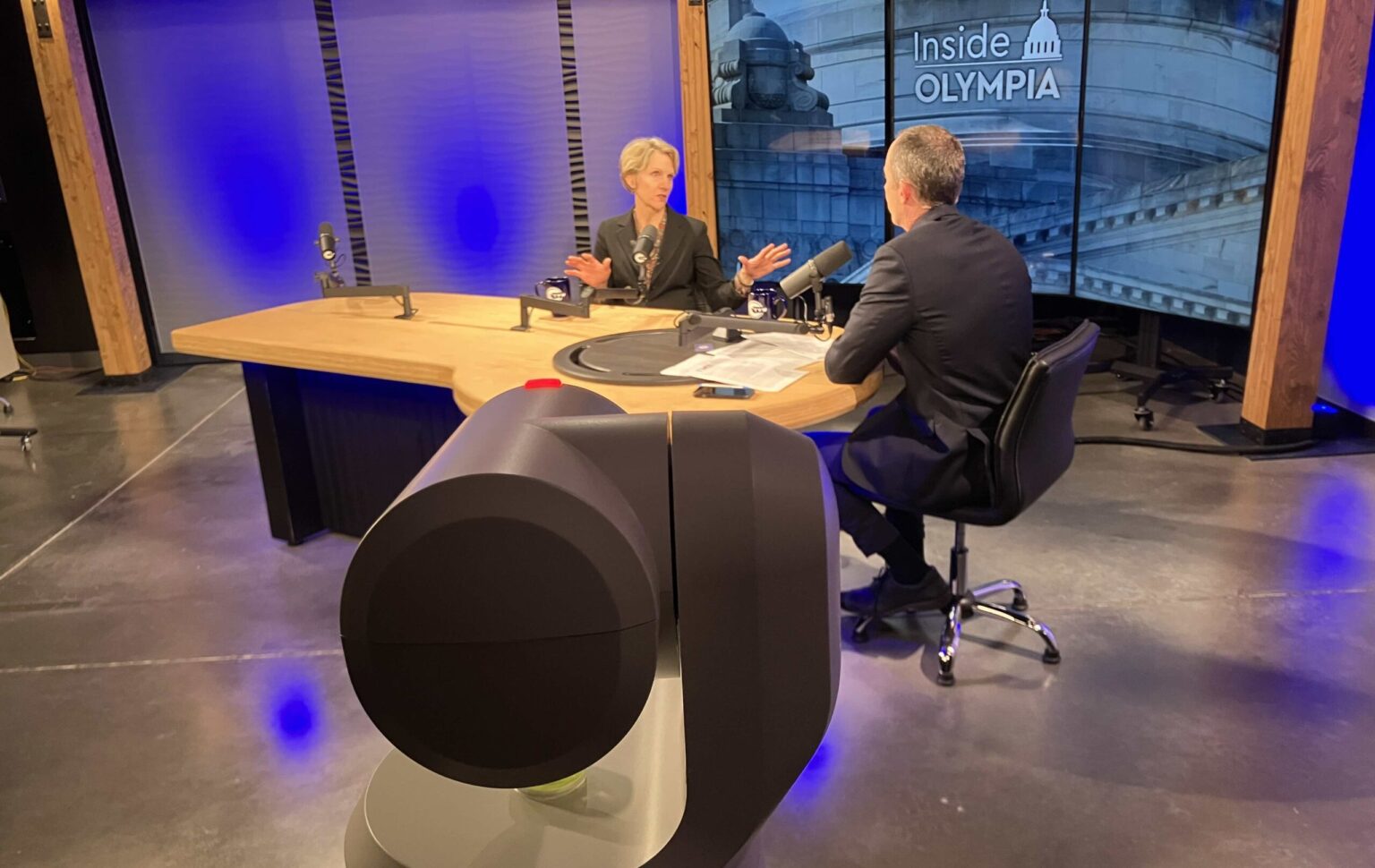 A television host seated in a studio during a political interview, looking down at notes, news photography style