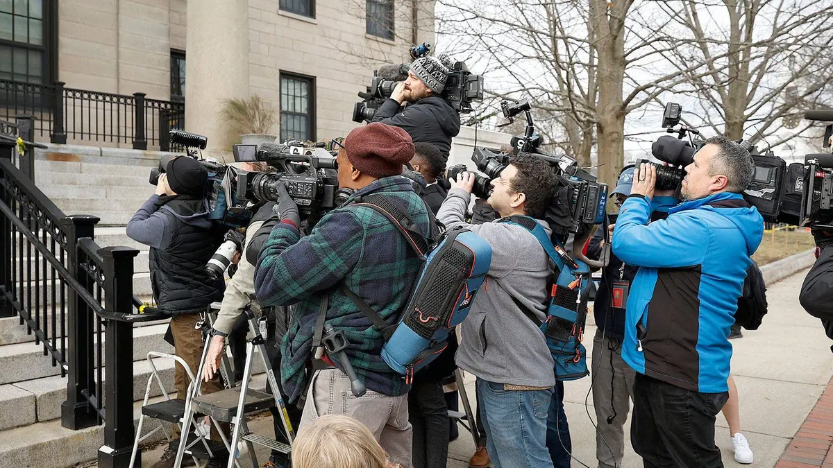 A television news camera crew set up on a sidewalk outside a courthouse entrance, with reporters waiting in the background, news photography style