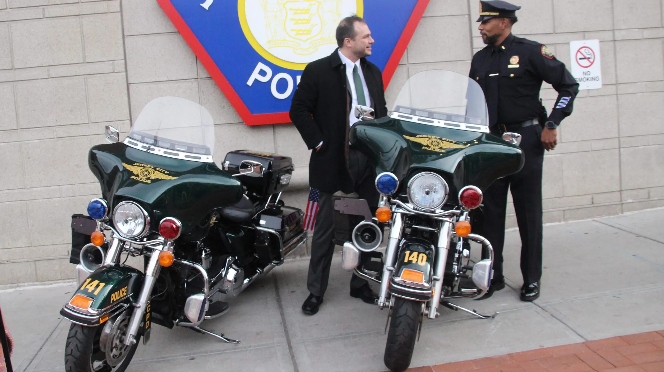 A uniformed Jersey City police officer standing beside a marked patrol car near a city intersection in daylight, news photography style
