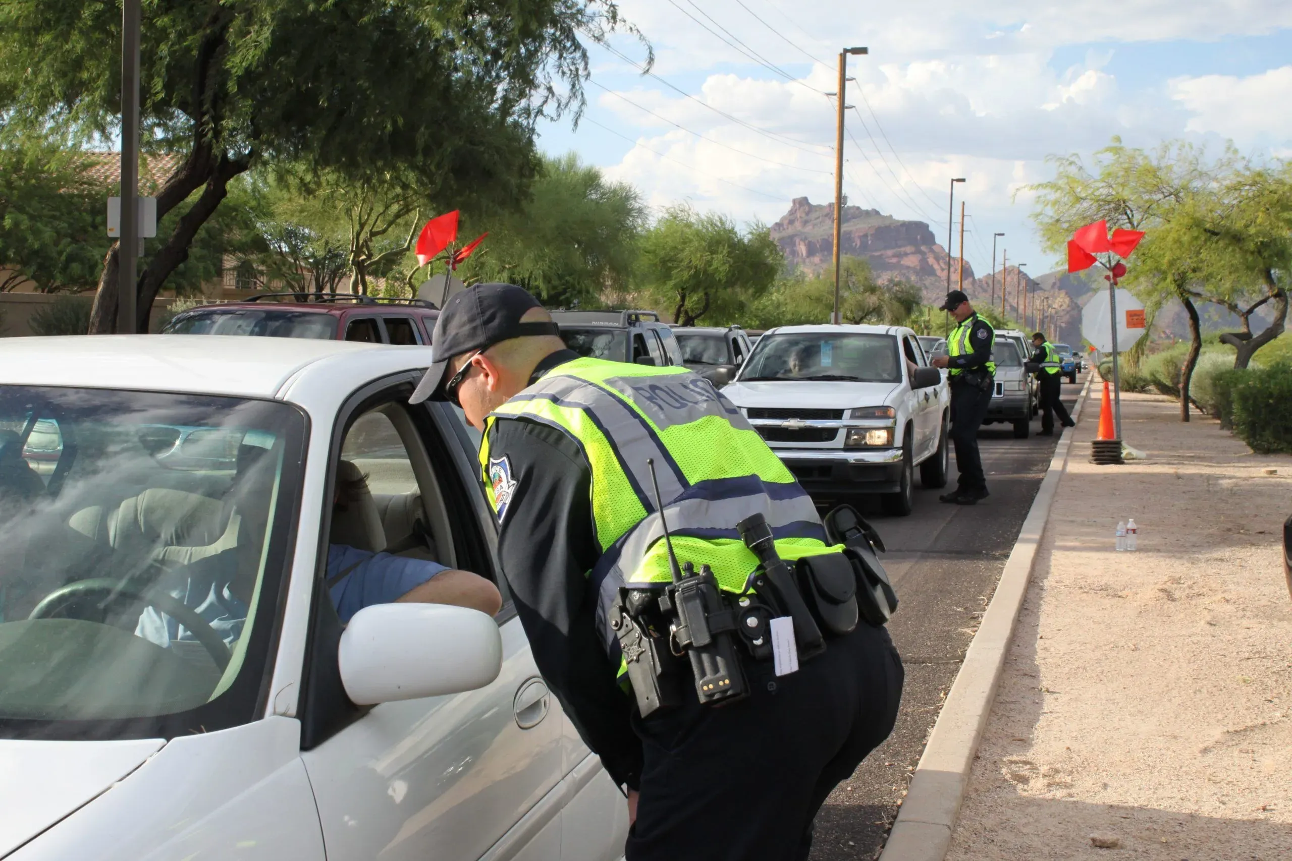 A uniformed police officer standing beside a stopped sedan at a county road sobriety checkpoint at dusk, with traffic cones and a patrol SUV visible in the background, news photography style