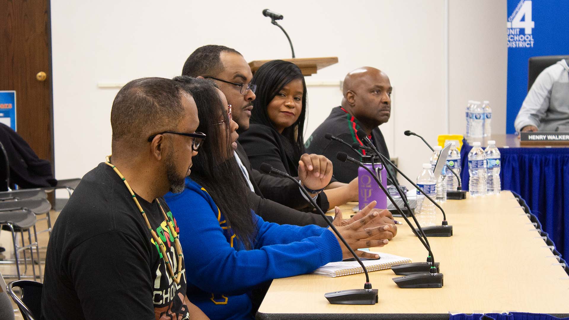 A university administrator speaking at a podium during a campus press conference with microphones and a campus building in the background, news photography style