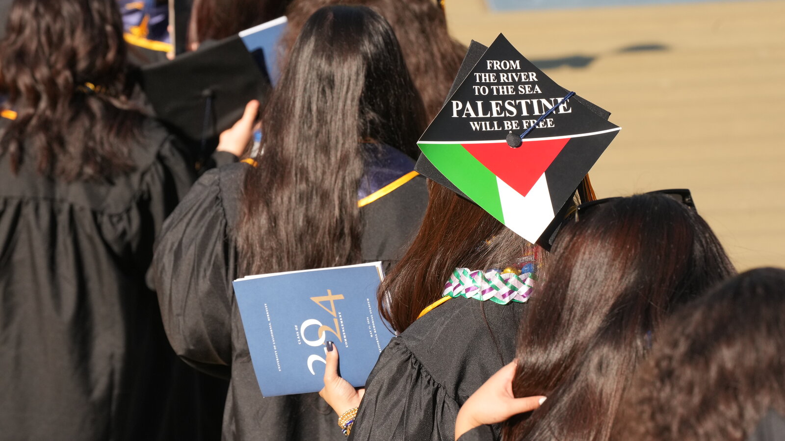A university commencement ceremony with graduates in caps and gowns as a small group holds political signs near the edge of the crowd, news photography style