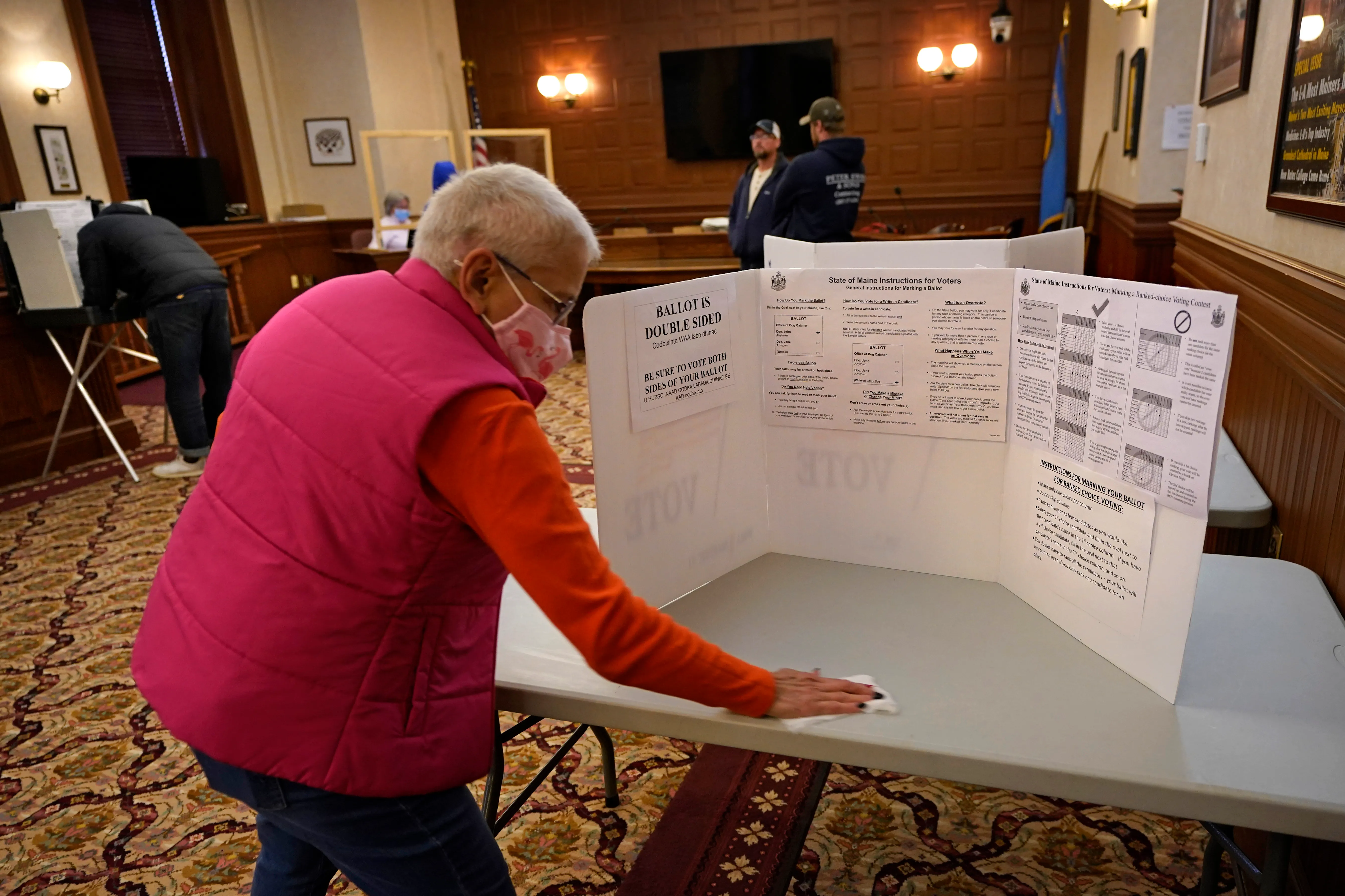 A voter standing at a privacy booth in a local election polling place, holding a paper ballot and filling in ranked-choice ovals with a pen, documentary news photo style