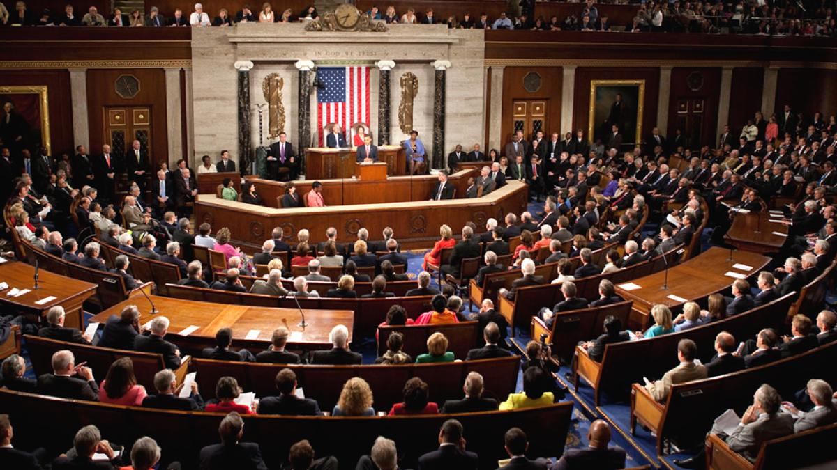 A wide angle real photograph of the United States House of Representatives chamber during a vote, with members seated and standing while the rostrum and clerk area are visible