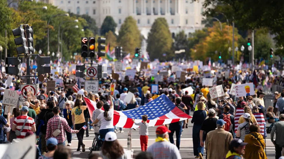 A wide street-level photograph of a peaceful crowd at a daytime protest holding handmade signs and American flags, with a city skyline in the distance, documentary news photography