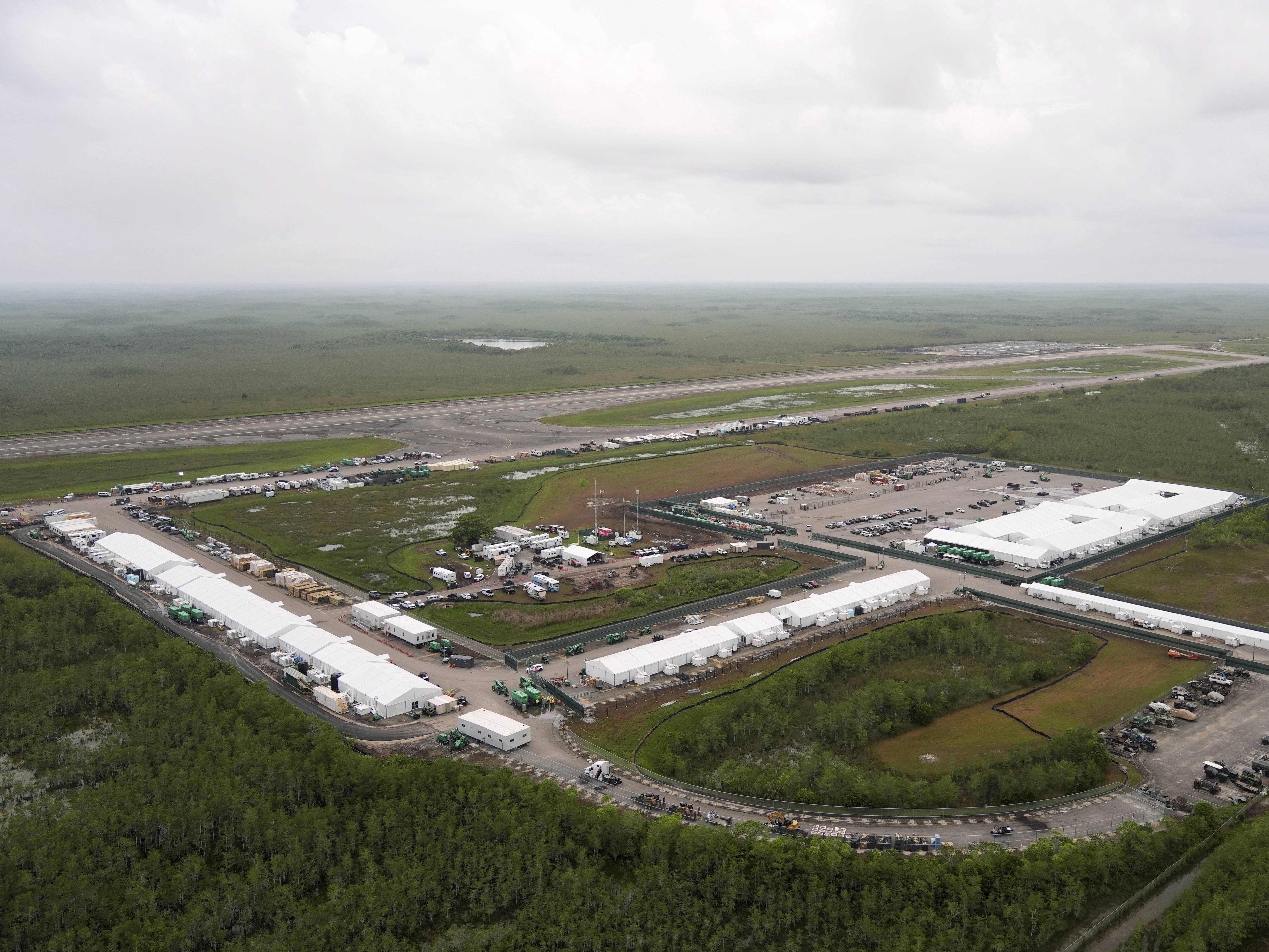 A wide view of a fenced perimeter at an immigration detention facility near the Florida Everglades, with flat wetlands in the background, news photography style