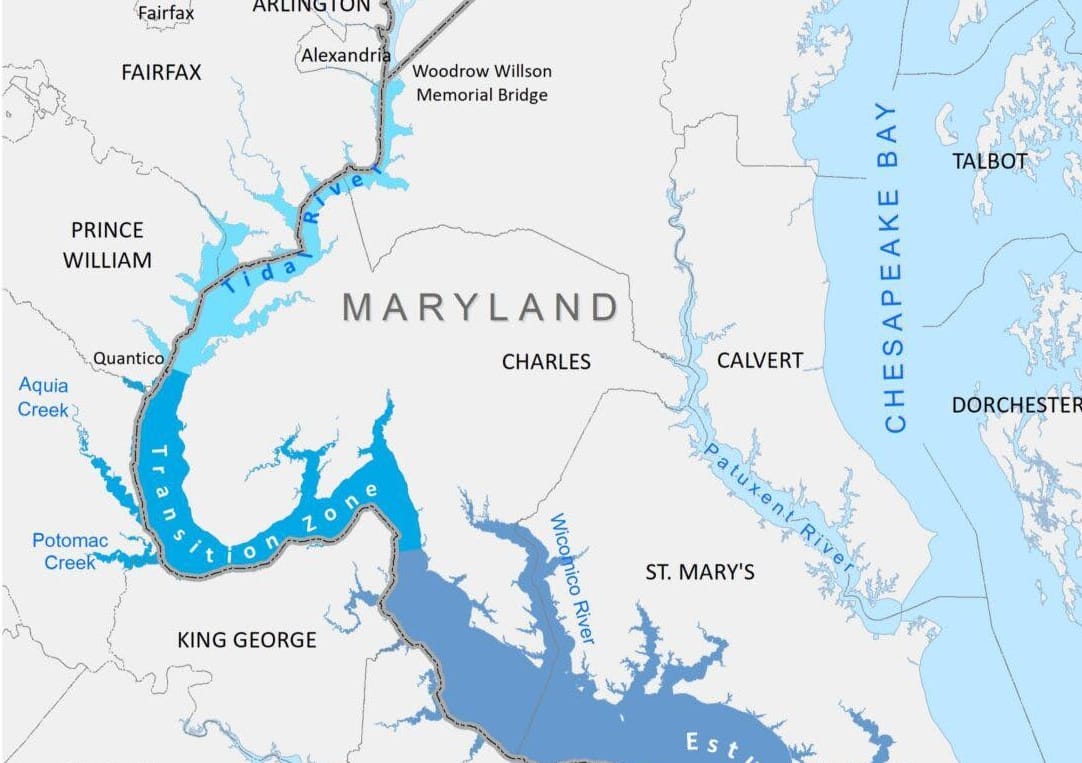 A wide view of the Potomac River from the shoreline with calm water and wooded banks in the distance, photographed in natural light, news photography style