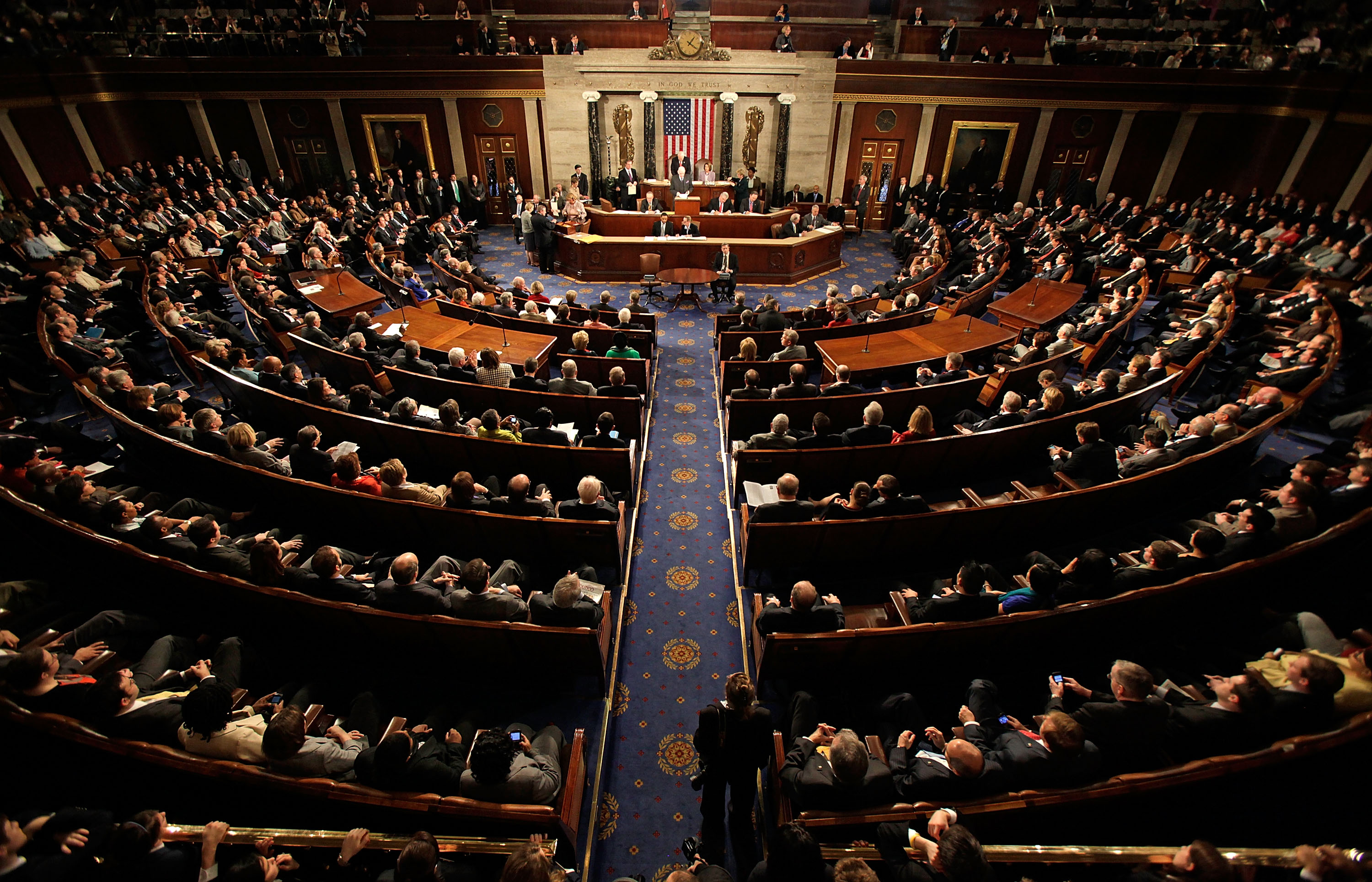 A wide view of the U.S. House chamber during a session at the Capitol, with lawmakers seated and staff moving through the aisles, photojournalism style