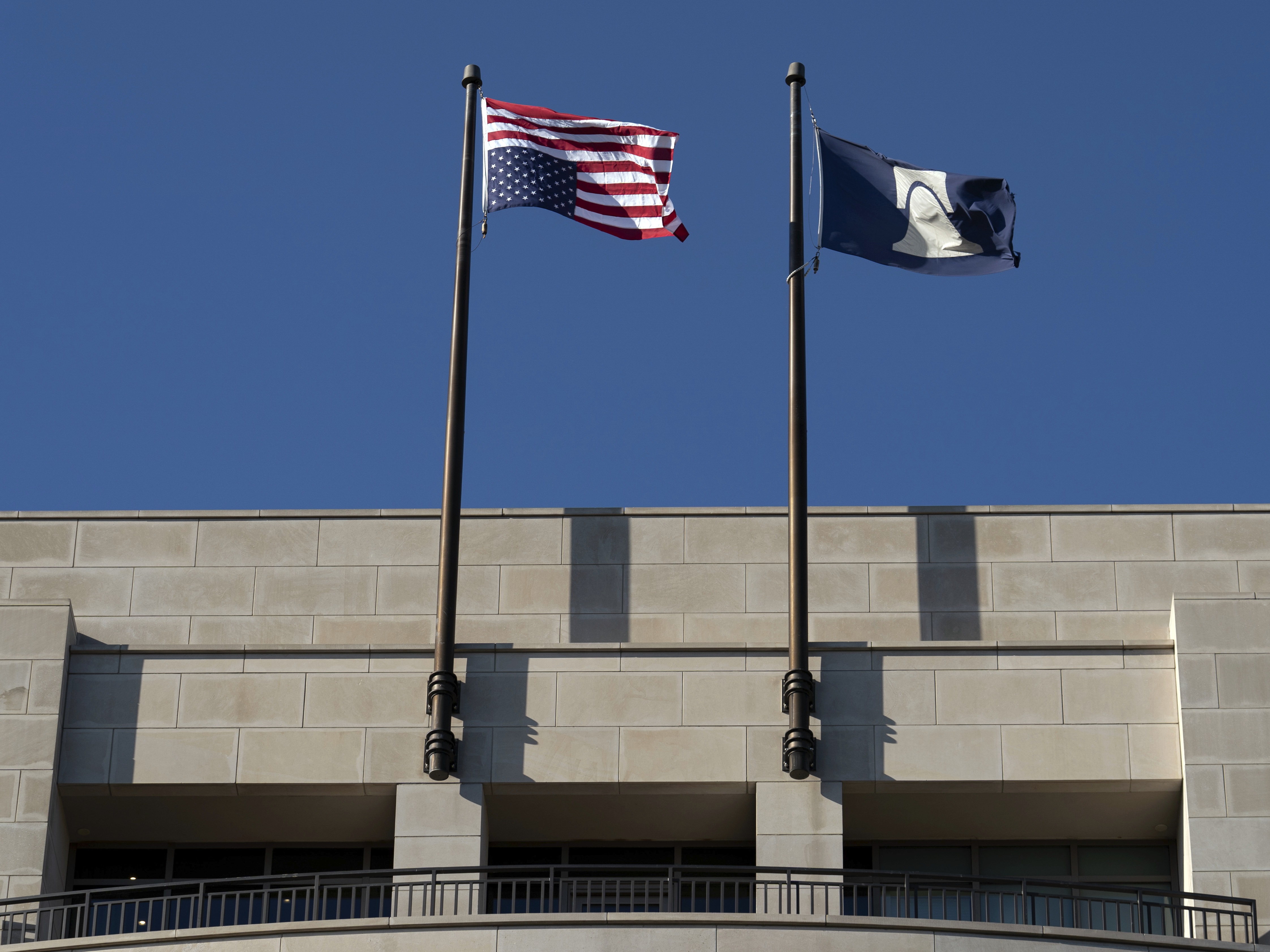 An American flag waving on a pole outside a federal courthouse plaza on a breezy day, with pedestrians walking in the background, news photography style