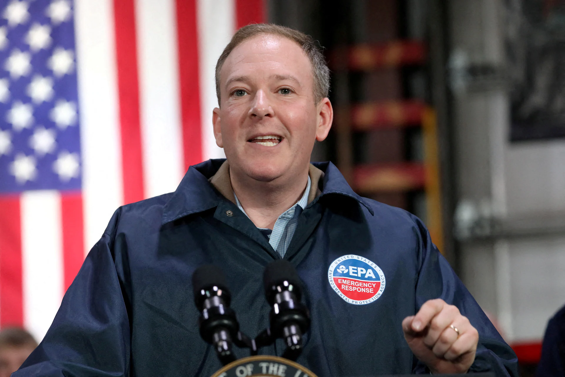 An EPA official speaking at a press conference podium indoors with reporters seated in the foreground, microphones visible, realistic news photography