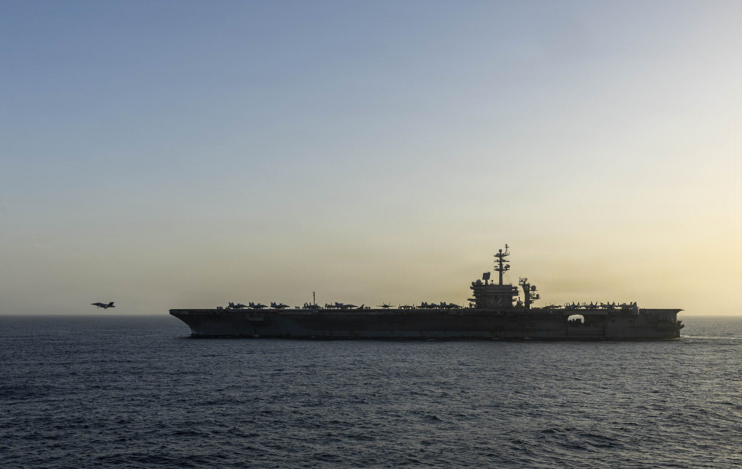 An Iranian-flagged cargo vessel moving through the Strait of Hormuz with naval ships nearby on the horizon, news photography style