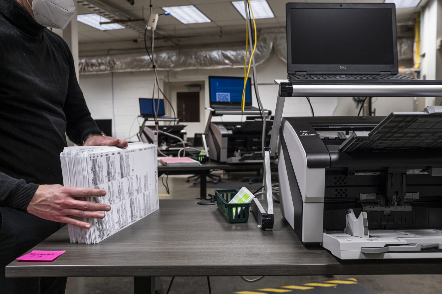 An election worker standing beside a ballot printing machine in a county election office, with stacks of paper in the background, news photography style