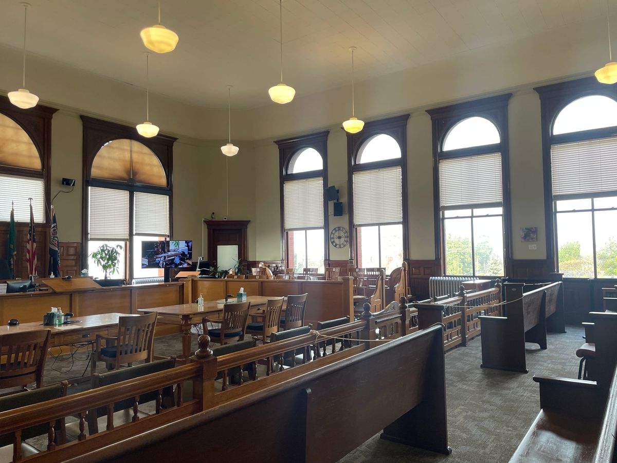 An empty witness stand and judge’s bench inside a quiet courtroom with wooden paneling, photographed from the gallery in natural indoor light