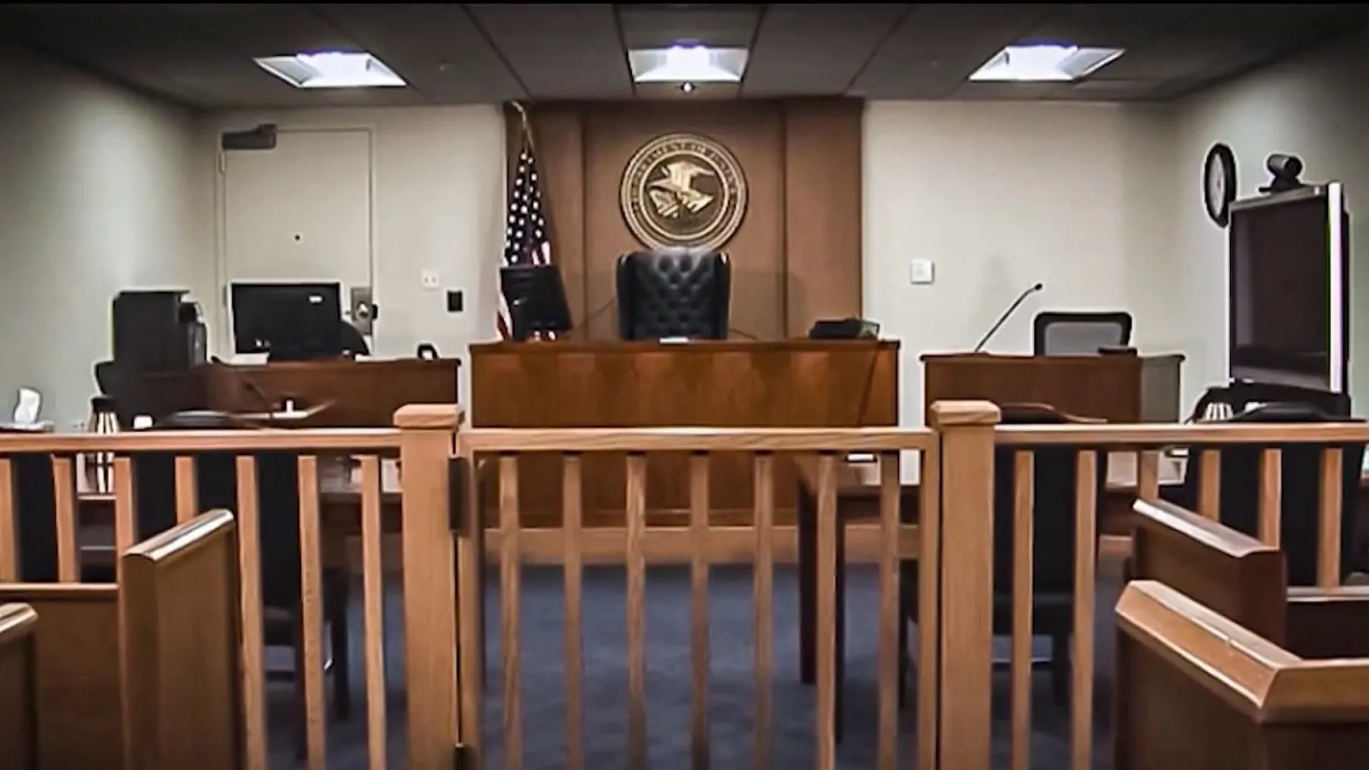 An immigration courtroom interior showing the judge’s bench, the government counsel table, and the respondent’s table, photographed from the public seating area, realistic news photography style