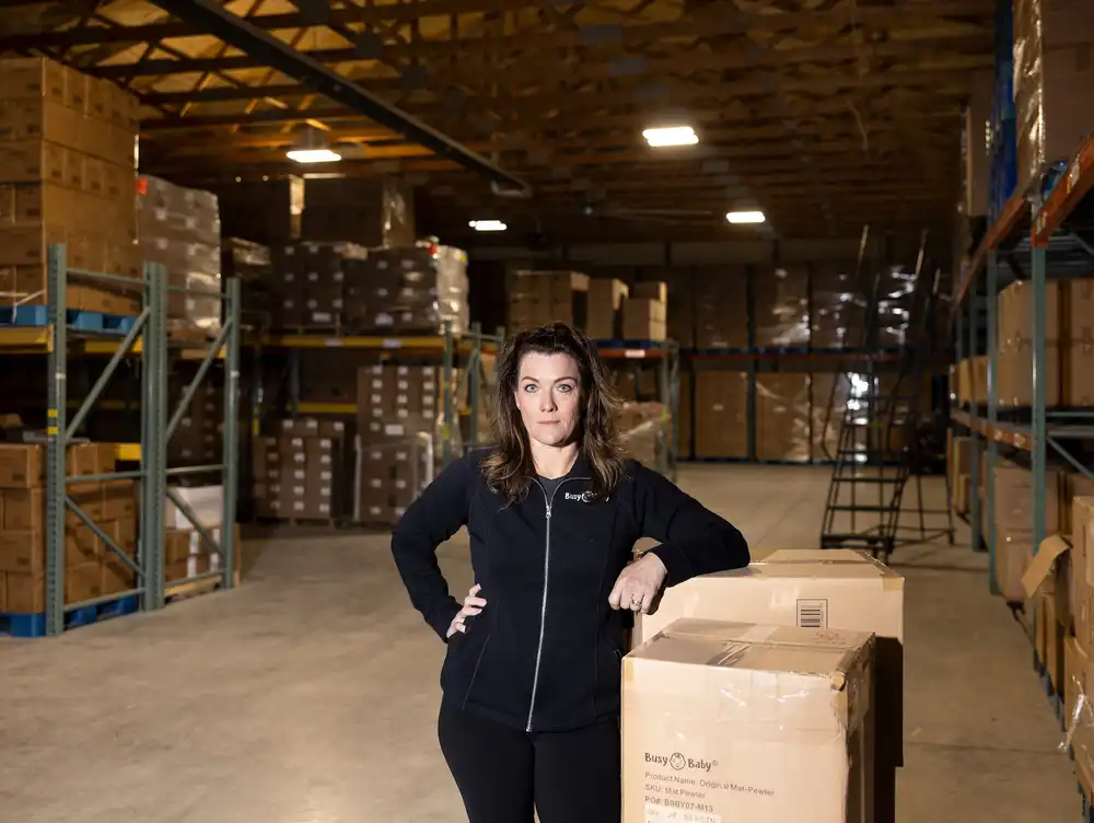 Beth Benike of Busy Baby standing inside a small warehouse with shelves of packaged baby products behind her, candid news photography style
