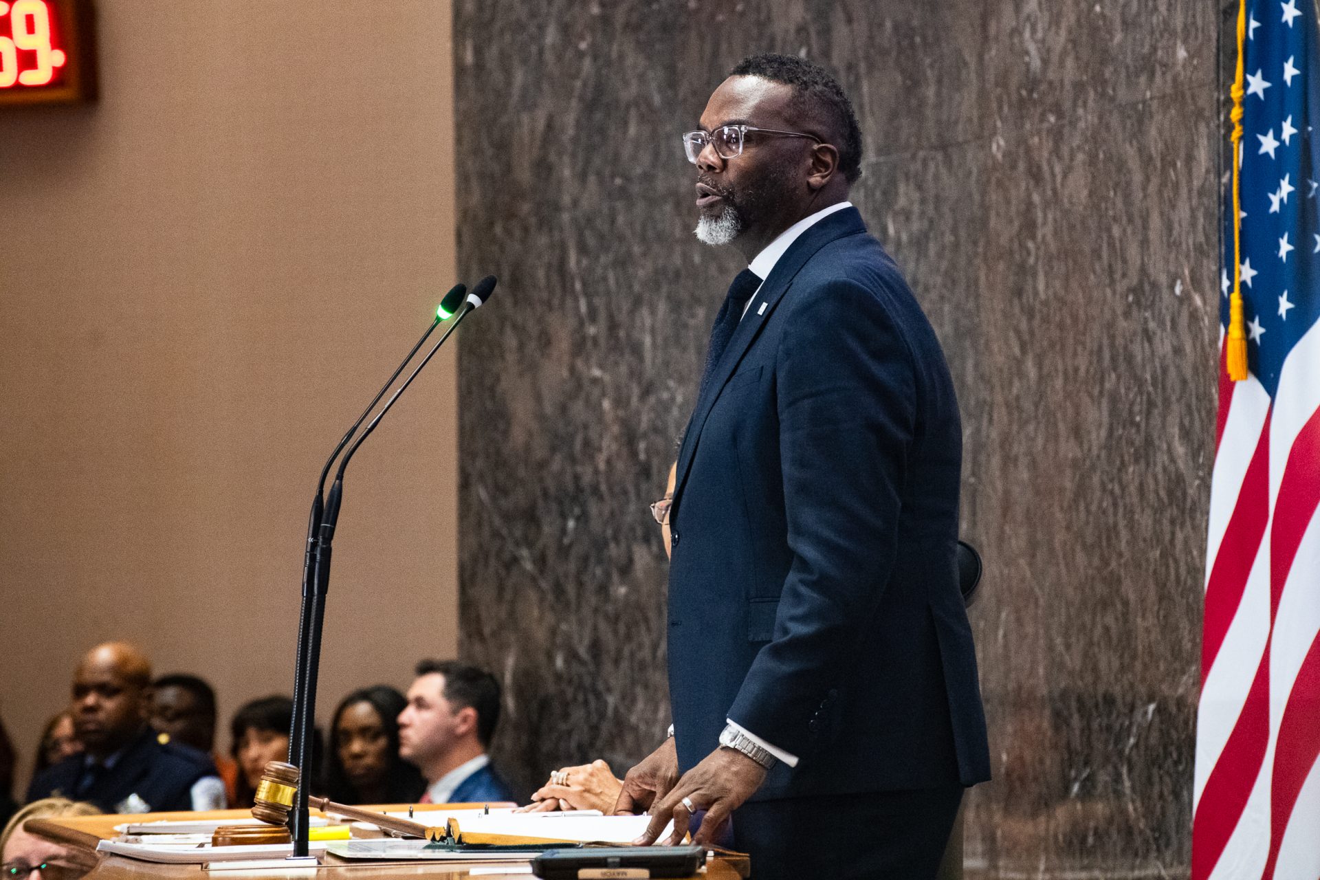 Brandon Johnson seated at a Chicago City Hall meeting table with microphones and officials in the background, news photography style