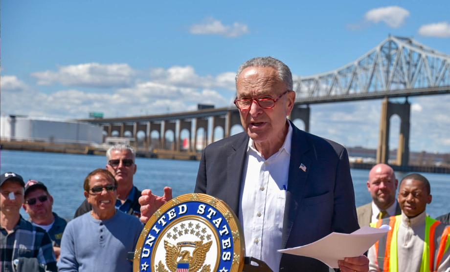Chuck Schumer speaking at a Senate press availability in the U.S. Capitol hallway with reporters and microphones in the foreground, photorealistic news photography