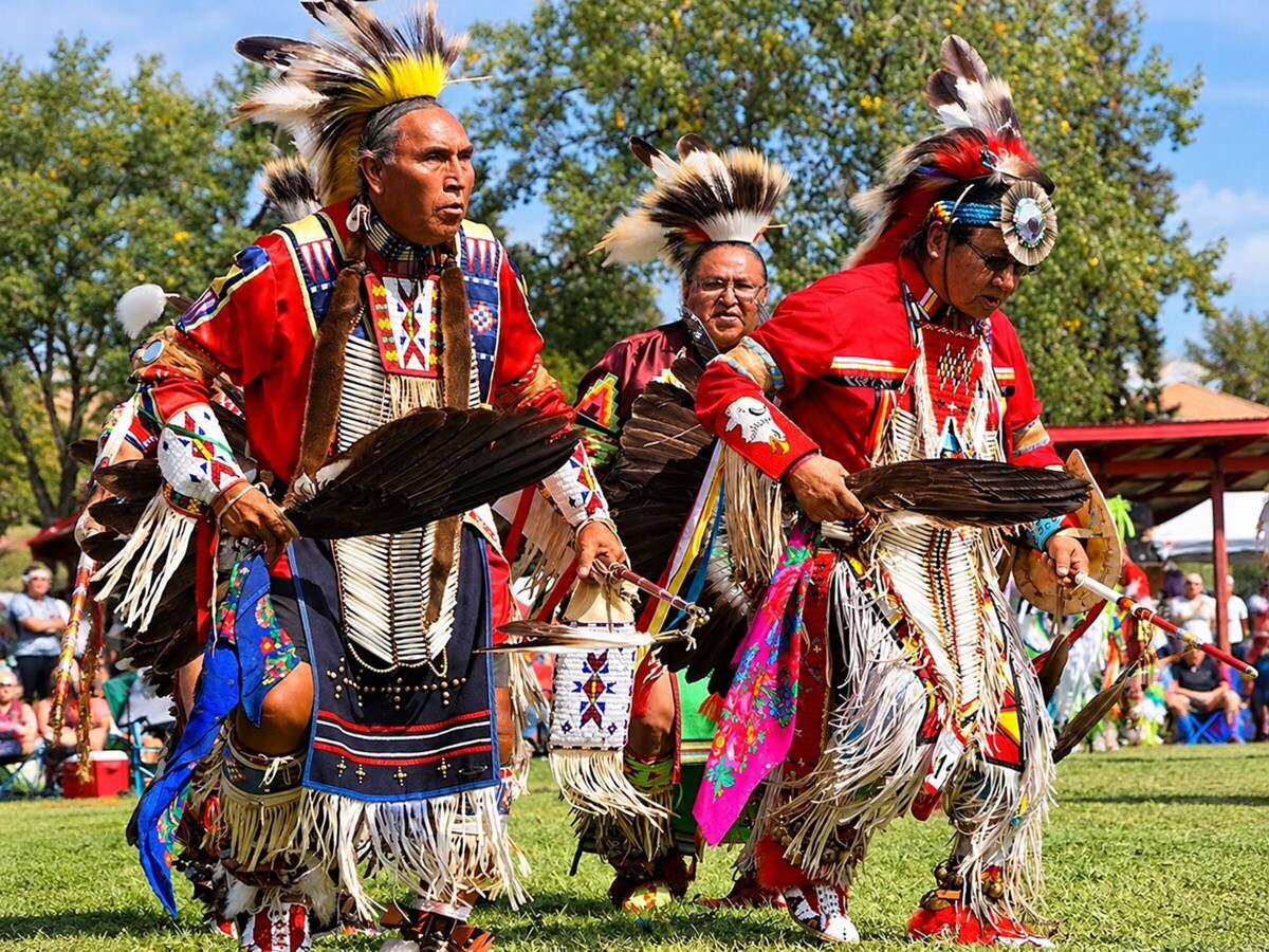 Community gathering featuring Indigenous dancers in regalia performing outdoors, with informational booths and banners referencing Indigenous heritage and resilience.