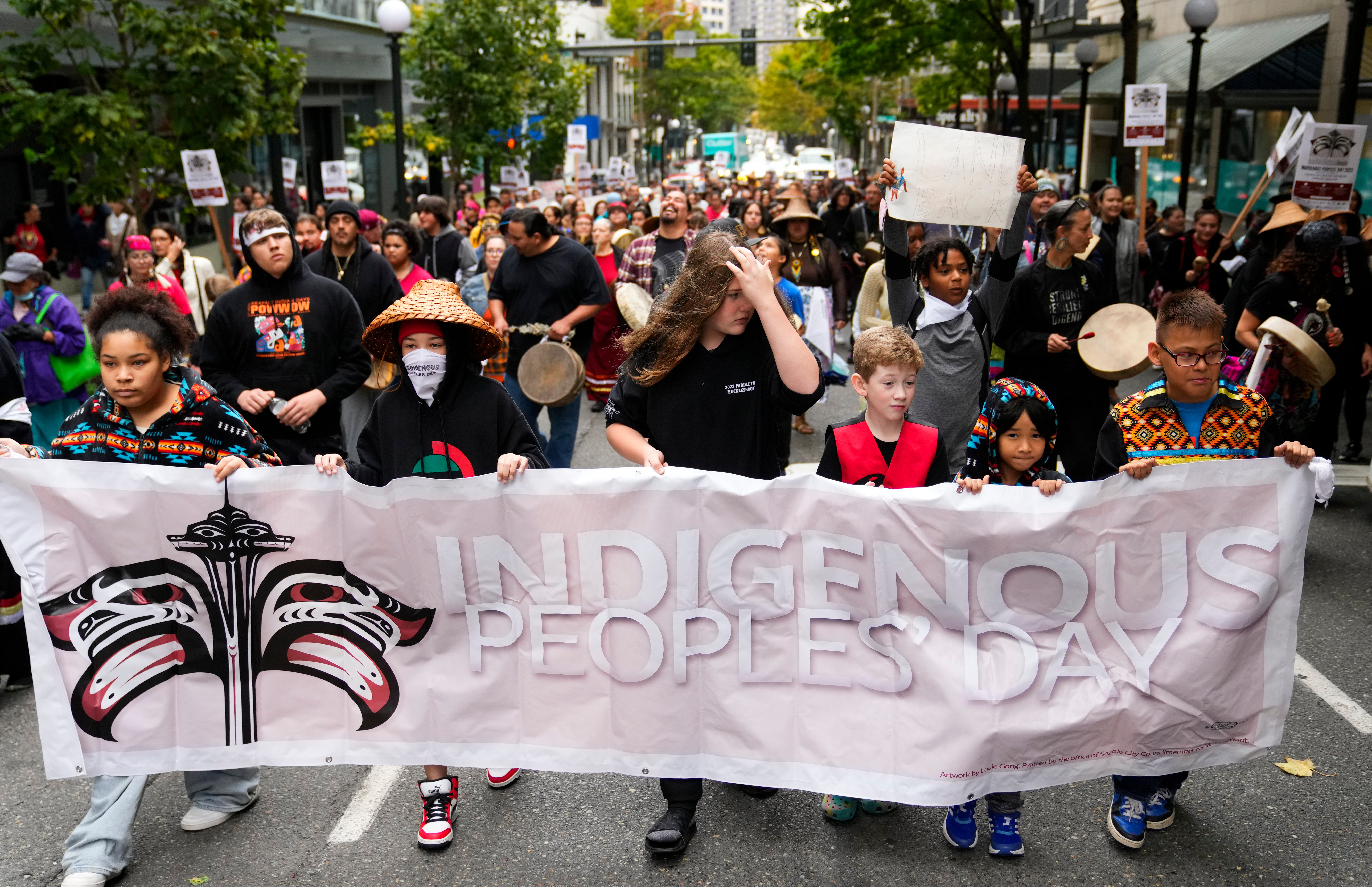 Crowd at a civic rally holding clear “Indigenous Peoples’ Day” signs outside a government building, with diverse ages and readable poster text.