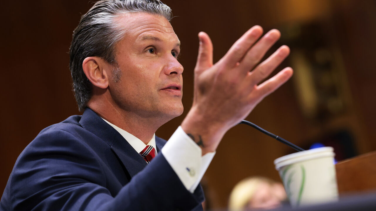 Defense Secretary Pete Hegseth seated at a witness table during a U.S. Senate hearing, speaking into a microphone with senators visible in the background