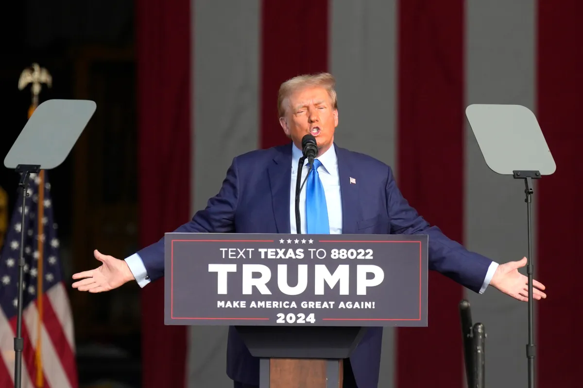 Donald Trump at a rally podium mid-speech with supporters and American flags in the background, telephoto news photograph