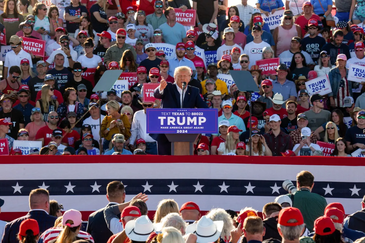 Donald Trump speaking at a Texas campaign rally, standing at a podium under bright stage lighting with supporters and American flags in the background, news photography style