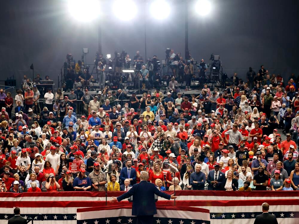 Donald Trump speaking at a campaign-style public event with American flags visible