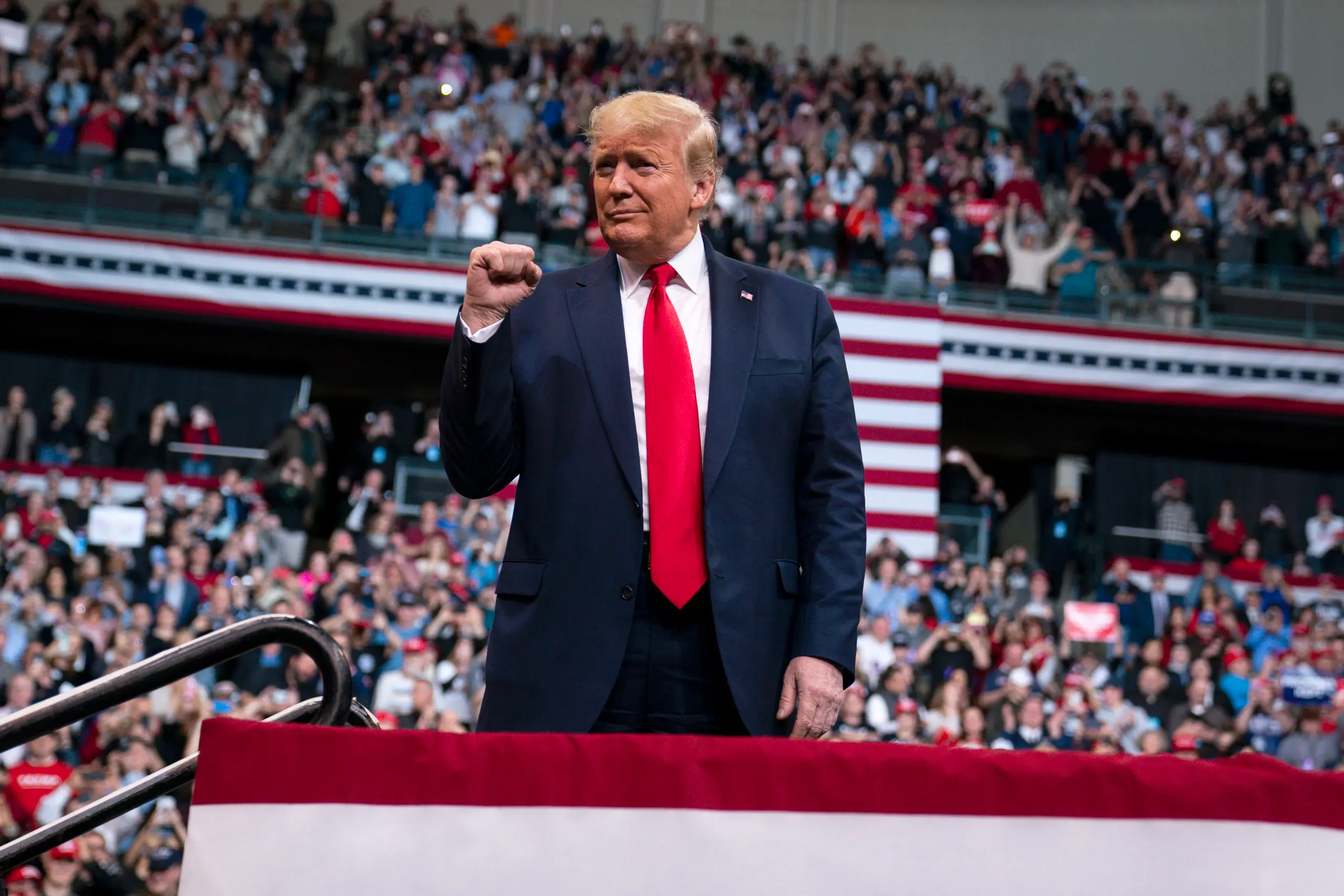 Donald Trump speaking at a public event, photographed at a lectern with microphones and a crowd in the background